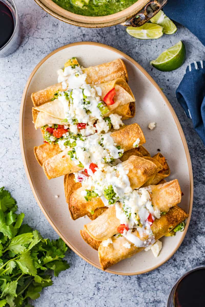 overhead image of chicken taquitos on a serving plate topped with crema, guacamole, and diced tomatoes